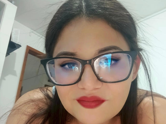 Nymphonebula23, com seios grandes, usando óculos pretos, batom vermelho e sombra azul, tem óculos refletivos que mostram seu reflexo, cabelo castanho e um tom de pele claro..