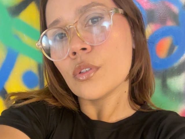 SophiaQueens with shiny lips, wearing clear glasses, black shirt, brown hair, and a slight pout, looking directly at the camera.