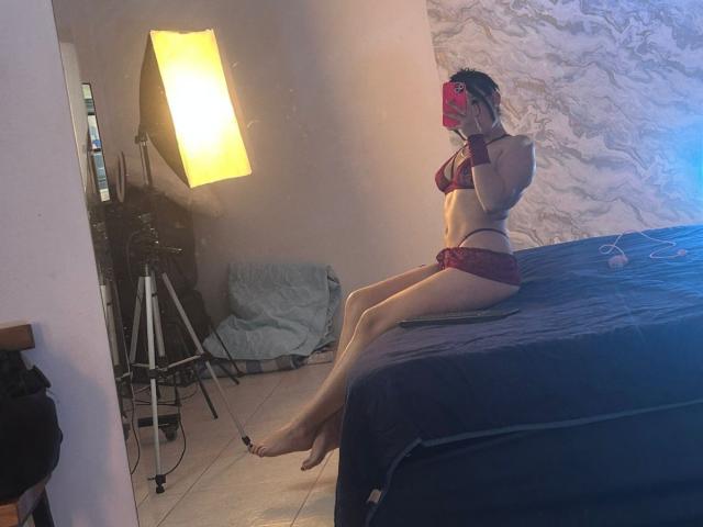TanyaEmas in red lingerie, sitting on a bed, showing her tits and ass, taking a mirror selfie with a pink phone, in a room with a standing light.