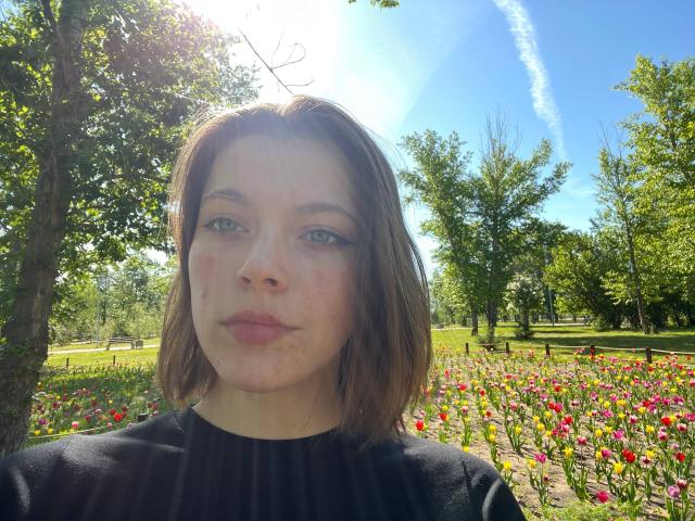 JennellMatacale has pale skin, brown shoulder-length hair, blue eyes, and slight freckles, wearing a black top, standing in a sunlit flower field.