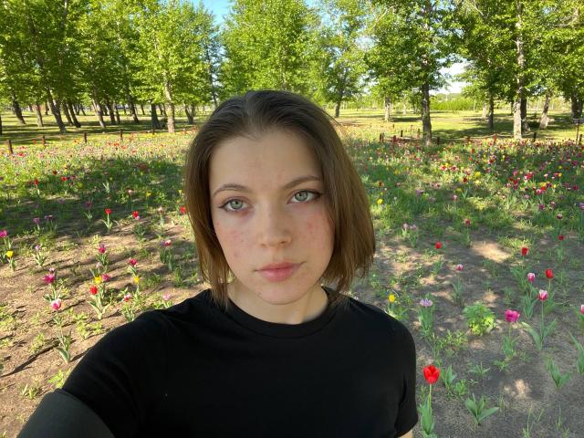JennellMatacale with short brown hair, green eyes, and light freckles, wearing a black shirt, standing in a sunlit flower field. Boobs not visible. Ass not visible. Pussy not visible.