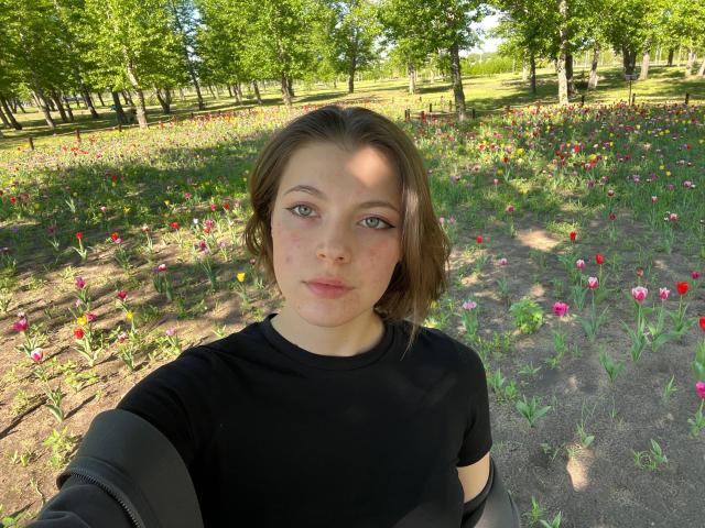 JennellMatacale has short brown hair, green eyes, and a slightly flushed face, wearing a black shirt, standing in a sunlit flower field with red and pink tulips.