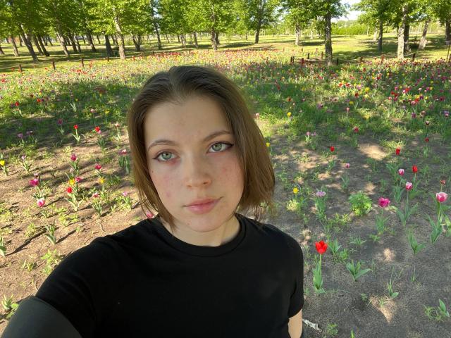 JennellMatacale with short brown hair, green eyes, slight freckles, black shirt, standing in a sunlit flower field, looking at the camera with a neutral expression.