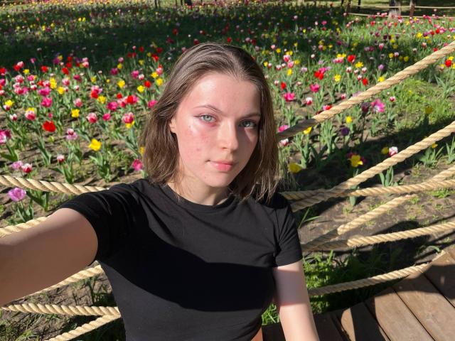 JennellMatacale with fair skin, green eyes, and pink cheekmarks, wearing a black shirt, taking a selfie in a sunlit flower field with colorful tulips and rope fencing.