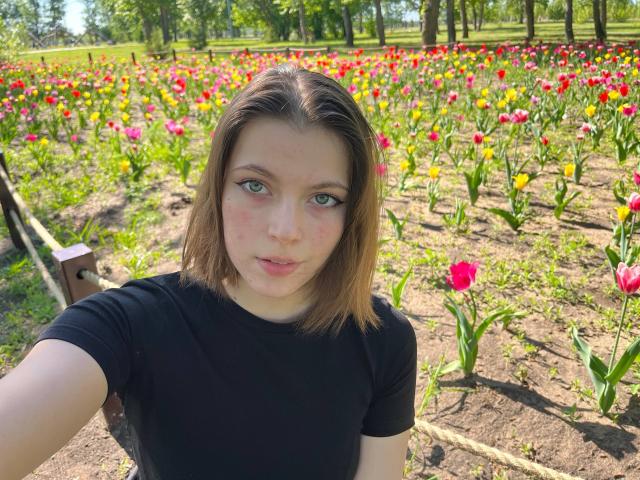 JennellMatacale with short brown hair, black t-shirt, green eyes, and freckles, standing in a sunny flower field, surrounded by colorful tulips. Boobs are not visible.
