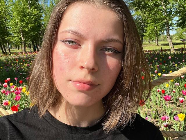 JennellMatacale has a shiny, pinkish face with slight acne, light brown shoulder-length hair, green eyes, and pinkish lips. She's wearing a black shirt in a sunny flower field.