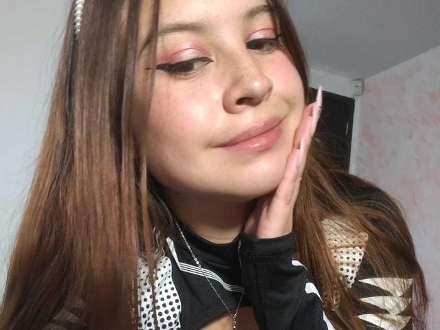 Melani Becker a des fards à paupières roses, de longs cheveux bruns et un serre-tête en perles. Elle sourit, sa main sur sa joue, et montre son haut noir et blanc..