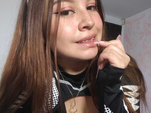 Melani Becker, avec ses longs cheveux bruns, son fard à paupières rose et un léger sourire, mordille son doigt, portant un haut noir et blanc à motifs, qui dévoile sa poitrine et où elle porte un collier..