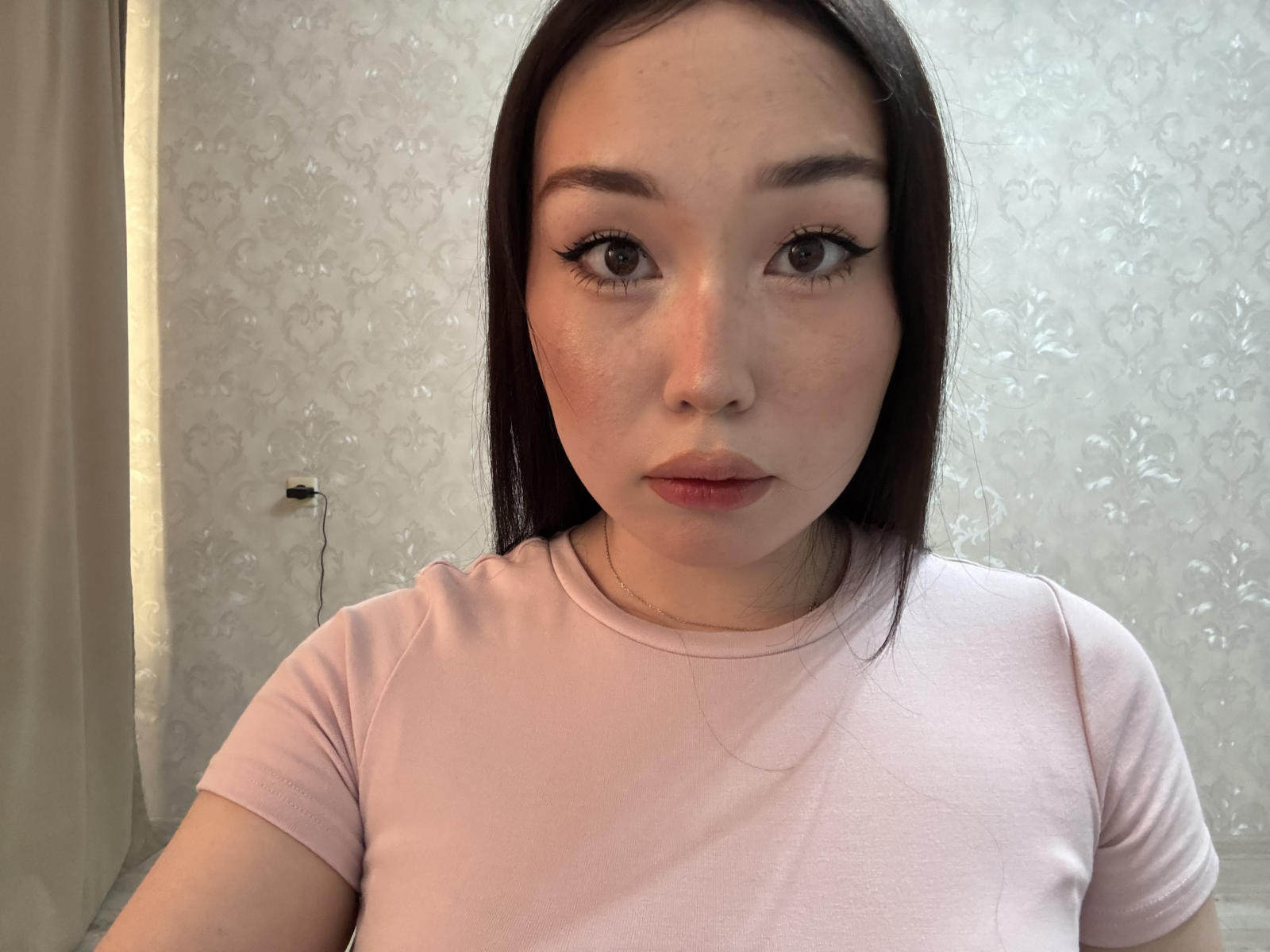 Lorenza Mccaughan, con lunghi capelli scuri, indossa una camicia rosa, ha delle lentiggini, eyeliner e un'espressione neutra, mostrando il suo viso e la parte superiore del petto, in una stanza con una luce soffusa..