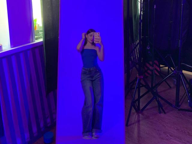 XeniaSherod with blue strapless top and high-waisted jeans, standing in front of a mirror, taking a selfie with hands in her brown hair, in a dimly lit room.