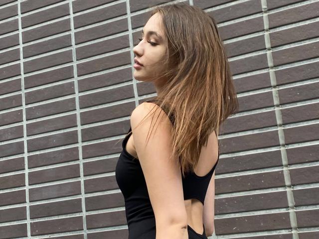XeniaSherod with her eyes closed, wearing a black cut-out tank top, showing her back and arms, brown hair, and a slightly open mouth. Background is a grid wall.
