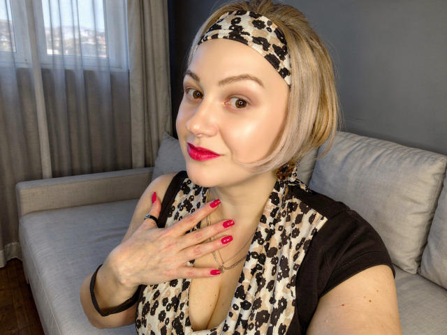 MissisXL com cabelo curto loiro, faixa de cabeça com estampa de leopardo, batom vermelho, blusa preta e cachecol com estampa de leopardo, exibindo unhas vermelhas e um piercing no nariz, sorrindo para a câmera..