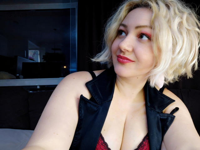 Una mujer rubia, MissisXL, con cabello corto y ondulado, delineador de ojos rojo y lápiz labial rosa, vestida con una blusa negra sin mangas y escotada, que deja ver sus pechos cubiertos con encaje rojo y negro. Sonríe suavemente..