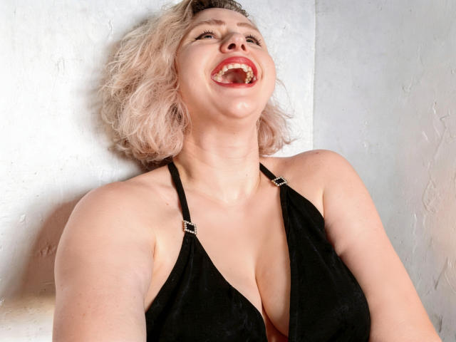 MissisXL mit blonden, lockigen Haaren, einem schwarzen Top mit Halter, und offenem Mund, der roten Lippenstift und ein leichtes Lächeln zeigt, hat große Brüste und helle Haut und lehnt sich an eine weiße Wand..