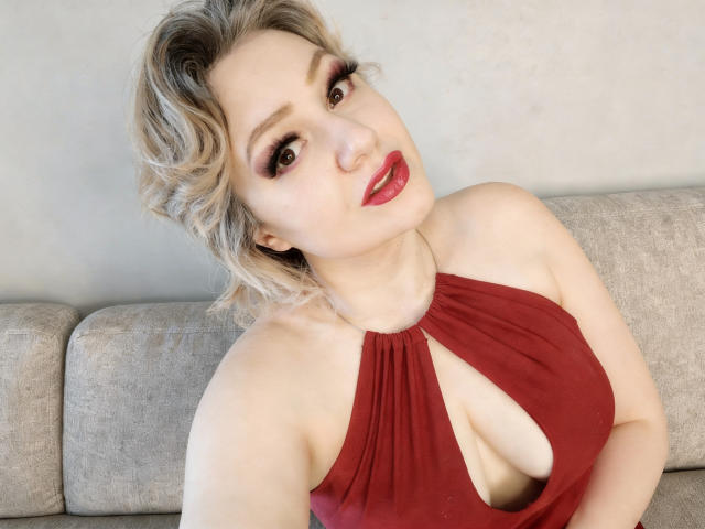 MissisXL with blonde, wavy hair wearing a red, halter dress with a deep cleavage, showing her large, round tits. She has red lipstick and is sitting on a beige couch.
