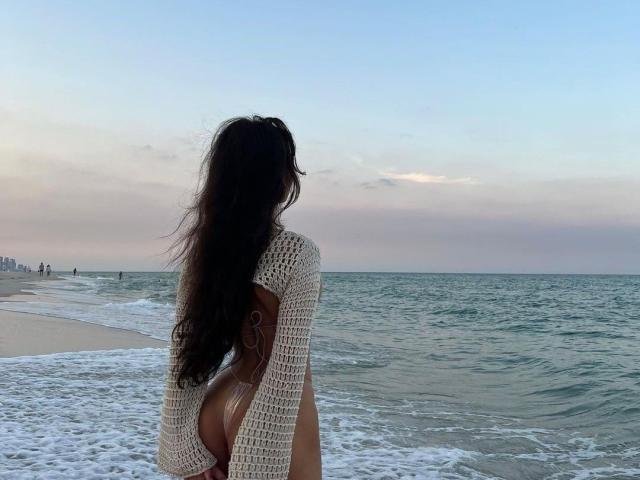 DianaFernandezz se tient sur la plage, dos à la caméra, vêtue d'un pull blanc en tricot qui dévoile ses fesses, avec de longs cheveux noirs et un ciel clair au coucher du soleil..