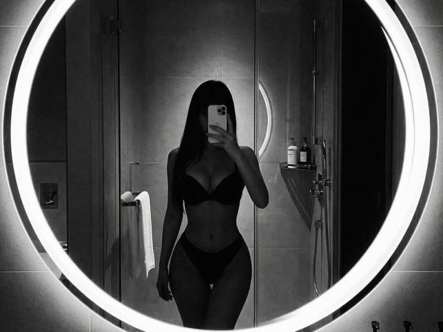 Autoportrait en noir et blanc de DianaFernandezz, avec de longs cheveux foncés, portant un soutien-gorge et des sous-vêtements noirs, tenant un téléphone, un miroir rond avec un anneau lumineux, des articles de toilette de salle de bain visibles, silhouette élancée..