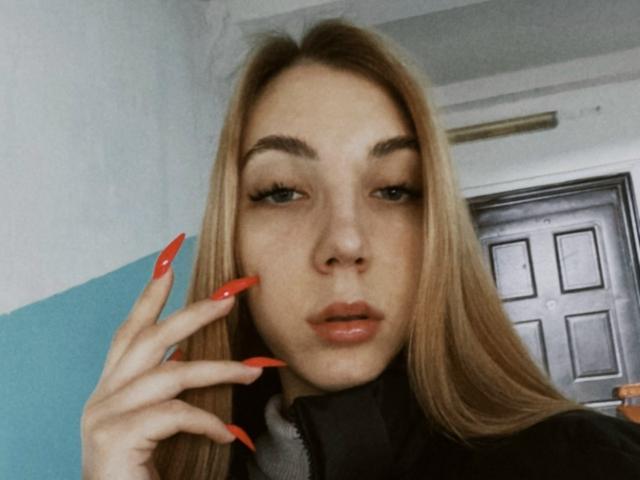 Alease Benson, avec de longs cheveux blonds, des ongles rouges, des lèvres pulpeuses et des sourcils épais, regarde l'appareil photo avec un léger moue, et porte une veste noire..