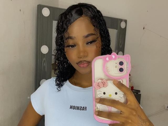 ShakiraThomps with curly black hair, wearing a white ribbed shirt, holding a pink Hello Kitty phone case, looking down at the camera with a neutral expression.