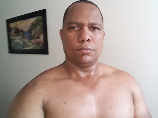 Shirtless BigBlackCock69, bald, medium brown skin, muscular chest, serious expression, small dark nipples, painted wall with blurred landscape in background.