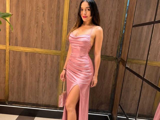 MindyLove in a pink, shiny, strapless dress with a high slit, showing her tits and legs, holding a pink purse, standing with red lipstick and long brown hair.