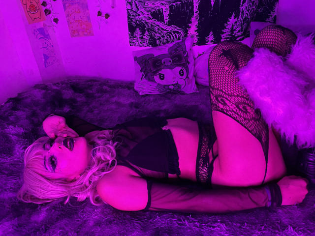 DollyBones with blonde hair, black lingerie, fishnet stockings, and thigh-highs, lying on purple-lit furry bed, legs up, white fluffy pillow, black bra, black panties, pink glow.