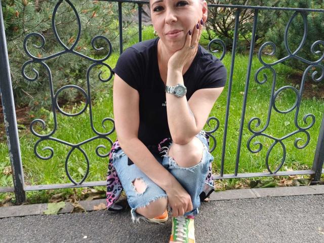 LauraSin, agachada con jeans rotos, una camiseta negra y zapatillas de colores, apoyando el mentón en la mano, llevando un reloj, con una valla de metal y césped verde detrás de ella..