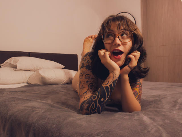 IsabellaArias with tattoos, lying on a bed, legs up, hands near mouth, glasses, black hair, surprised expression, grey blanket, white pillows, bare feet, wooden wall, red lipstick.