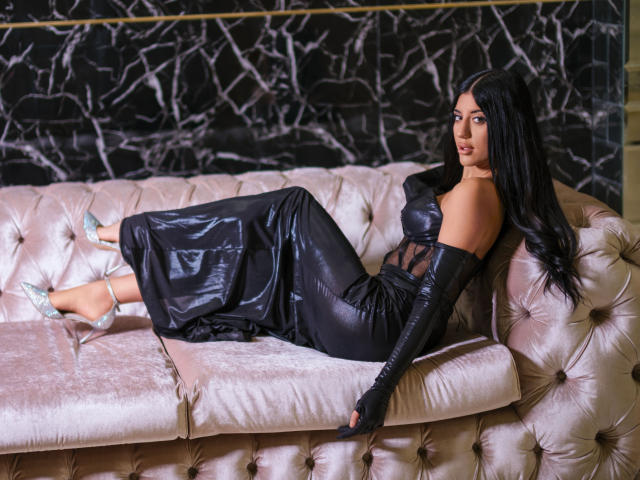 DariaVeil, vêtue d'une robe en cuir noir, avec de longs gants et des talons argentés, est allongée sur un canapé rose capitonné, montrant ses fesses et ses seins, avec des cheveux noirs et un regard séduisant..