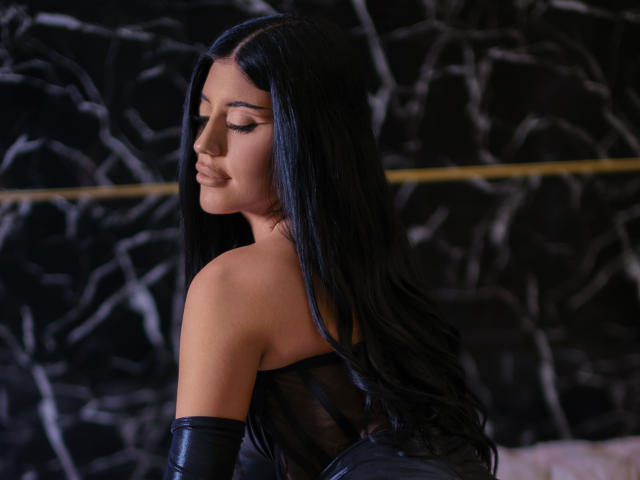 DariaVeil, avec de longs cheveux noirs, portant un haut noir transparent et une manche en cuir noir, regarde vers le bas avec une expression douce, ses fesses étant partiellement visibles..