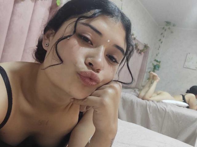 AmbarLandry with pouty lips, black hair, and small white dots on her forehead, wearing a black top, resting her chin on her hand, showing her tits and nose ring.