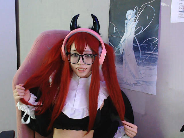 PaulineBitah con i capelli rossi, corna nere, occhiali, cuffie rosa, top nero, colletto bianco arricciato, che si tiene i capelli, mostra i denti, seduta su una sedia rosa, accanto a un poster anime..
