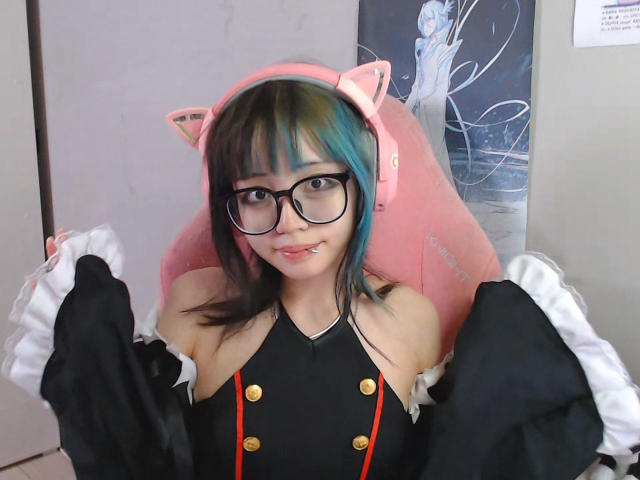 PaulineBitah avec des oreilles de chat, des cheveux noirs et bleus, des lunettes, une robe noire avec des boutons dorés, des volants blancs, et une chaise rose, souriante, montrant ses mamelons gonflés..