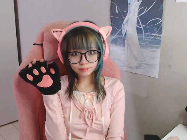 PaulineBitah, con gafas, orejas de gato rosas y guantes negros con forma de pata, sentada en una silla rosa, vistiendo una camisa rosa y una blusa blanca, mostrando una cara linda y ligeramente "nerd"..