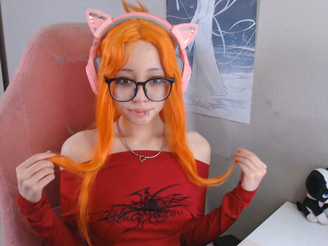 PaulineBitah, com cabelo laranja, orelhas de gato, óculos e piercing no nariz, segurando o cabelo, vestindo uma camisa vermelha com um ombro de fora, sentada em uma cadeira rosa. Seios visíveis sob a camisa..