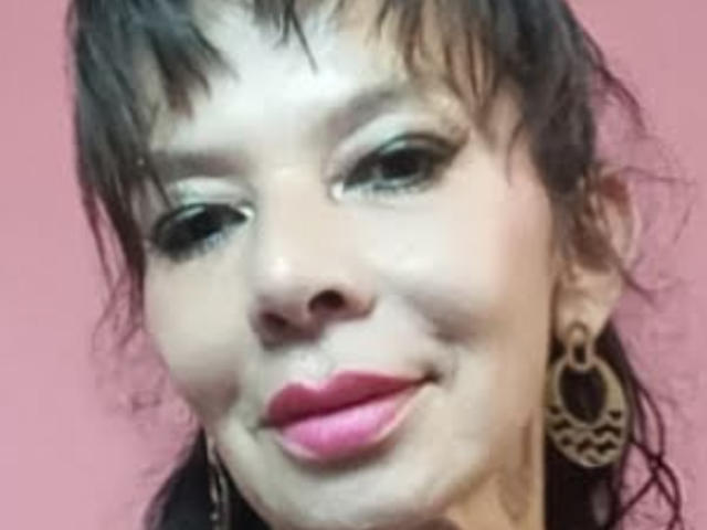 SamaraBellet hat braunes Haar, grüne Augen und rosafarbenen Lippenstift, und sie hat ein leichtes Lächeln. Sie trägt große, aufwendige Ohrringe und hat einen hellen Hautton. Ihr Gesicht ist leicht unscharf..