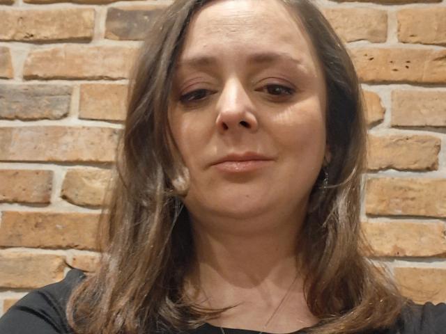 TaylorTrue tiene el cabello castaño, un tono de piel claro y una sonrisa sutil. Lleva una camisa negra y su nariz está ligeramente brillante. Sus senos no son visibles..