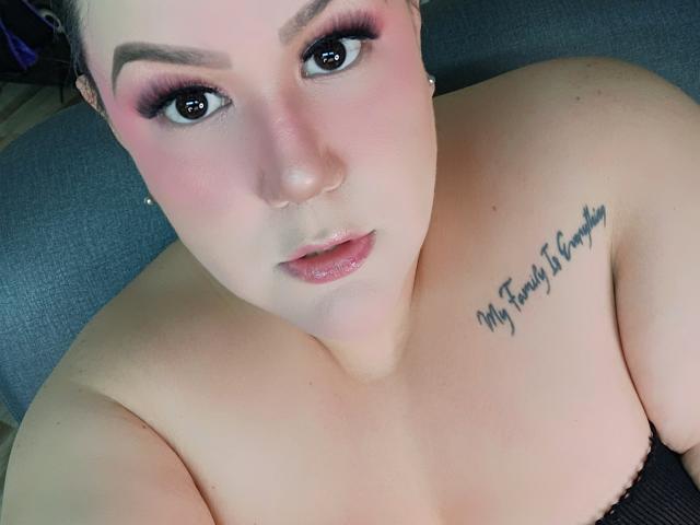 GiiannaMorrys mit rosigem Make-up, glänzenden Lippen und einem Tattoo auf ihrer Schulter, auf dem "Die einzige Firma meiner Familie" steht. Ihre Brüste sind freigelegt, und sie blickt nach oben in die Kamera..
