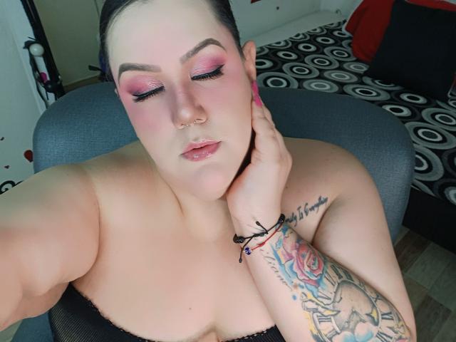 GiiannaMorrys with pink eyeshadow, nose ring, and tattooed arm, touching her neck, black strapless top, pink nails, and bracelets, in a gray chair, eyes closed.