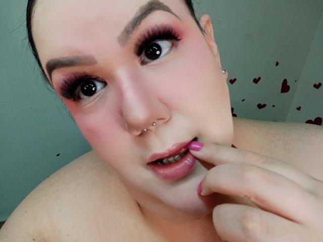 GiiannaMorrys con ombretto rosa, piercing al naso e rossetto rosa, che si morde un dito con le unghie rosa, guarda la fotocamera, mostrando le spalle scoperte e la pelle bianca..
