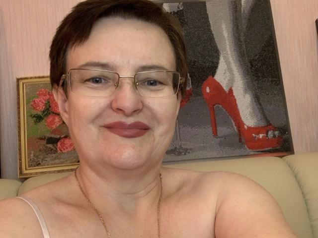 Margie Evans, con cabello castaño corto, gafas y una sonrisa discreta, lleva un collar dorado y un top sin tirantes que deja ver sus senos, en una foto donde también se ven unos tacones rojos..