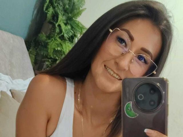 SharissWilliam with glasses, smiling, holding phone, white tank top, visible cleavage, brown hair, green eye makeup, gold necklace, green plant in background, selfie, phone case with green sticker.