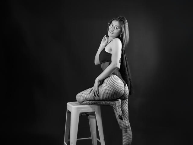 SharissWilliam with glasses, dark tank top, and fishnet pants, sitting on a stool, hand near mouth, looking back, black and white, showing her ass and a flirty smile.