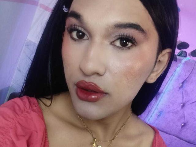 PamelaXFox has glossy red lips, dark straight hair, and a gold necklace with a small cross. She's wearing a pink top, looking at the camera with brown eyes.