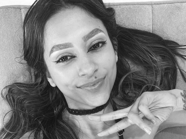 AfroditaWild, with long dark hair, thick eyebrows, and a nose ring, makes a peace sign with a black choker and eyeliner. Smiling, she has shiny lips and a cute face.