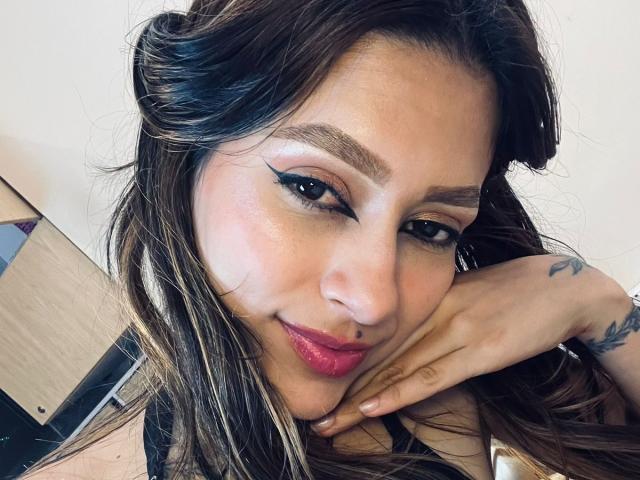 AfroditaWild has tan skin, dark brown wavy hair, bold eyeliner, pink lipstick, and a tattoo on her right hand, resting her cheek on her fingers, smiling.