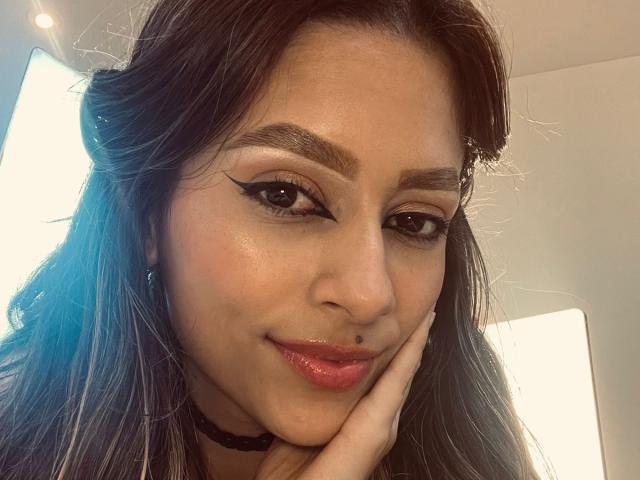 AfroditaWild with dark hair, black choker, winged eyeliner, and a small nose piercing, smiling while resting her cheek on her hand. Her glossy pink lips and brown eyes stand out.