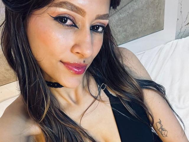 AfroditaWild with glossy pink lips, black eyeliner, and long brown hair, wearing a black top, showing a tattoo on her right arm, and a black choker.