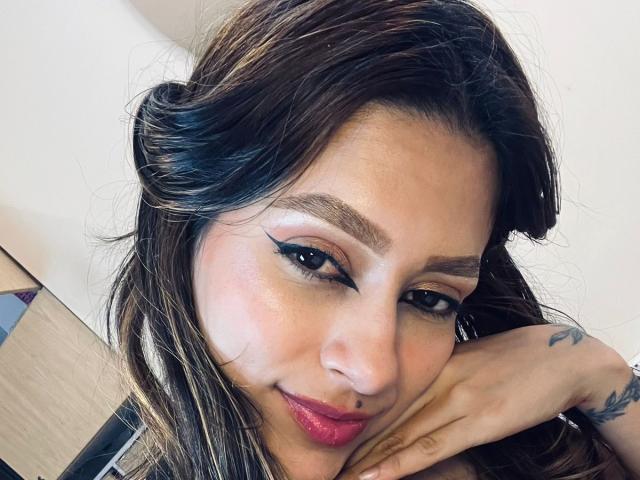 AfroditaWild has brown wavy hair, black eyeliner, pink lipstick, and a tattoo on her right hand. She's smiling with her chin resting on her hand.