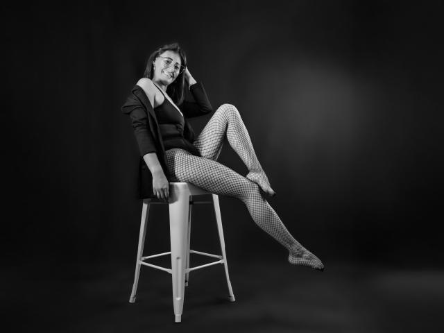 SharissWilliam is sitting on a stool, legs spread, wearing a black jacket, off-shoulder top, and fishnet stockings, with a relaxed, confident expression, and a slight smile.